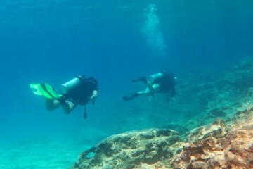 Wall Mural - Swimming scuba divers exploring shallow ocean, rocky seabed. Scuba divers in the clear water. Underwater photography from scuba diving. Seascape with rocks, travel photo. 