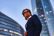 © Alex Vog - Perfect businessman in suit and sunglasses about skyscraper in business city district, serious looking away. Male person near build corporate company office in financial district. Copy ad text space