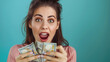 © Irina B - Young woman holding a stack of dollar bills, displaying excitement and joy against a teal background.