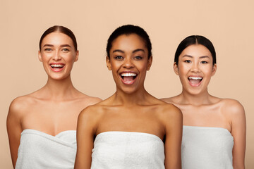 Wall Mural - Playful Multiethnic Model Girls Posing Wrapped In White Bath Towels Standing Over Gray Background, Smiling To Camera.