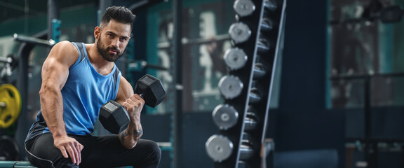 A man sits on a bench in a well-equipped gym, focusing on lifting a dumbbell with determination as he engages in his workout, panorama with copy space