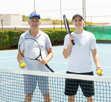 Smiling eldery man standing with son on outdoor tennis court.