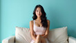 © DhanaStudio - Relaxed young Asian woman in casual wear sitting comfortably on light blue sofa, radiating tranquility and ease