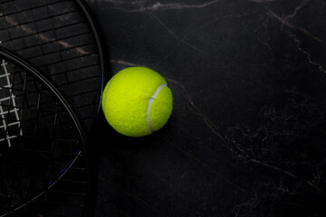 A yellow tennis ball lies against a background of black marble, dark tennis rackets are visible on the side. The concept of sports, competitions. High quality photo