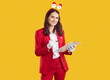 © Studio Romantic - Studio portrait of Santa female manager or event organizer with clipboard. Woman in red suit and funny Santa headband standing on yellow background and writing Christmas checklist on clip board