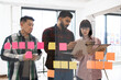 © sofiko14 - Group of young professionals collaborating on project in modern office. Team gathered in front of glass wall with colorful sticky notes while using digital devices for brainstorming and planning.