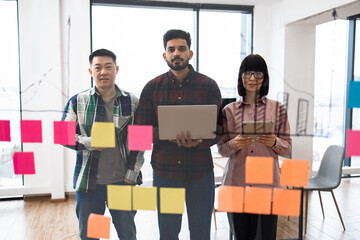 Wall Mural - Diverse team collaborating in modern office environment using sticky notes on glass wall. Team members with laptops and tablets planning and brainstorming. Modern workspace with city view.