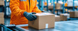 © Enjoy - A worker in an orange jacket carefully places package on conveyor belt in busy distribution center, ensuring quality control. environment is bustling with activity