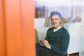 Wall Mural - Homeoffice for handsome man. Man sitting on floor and working on laptop. Through window shot. Telecommuting.