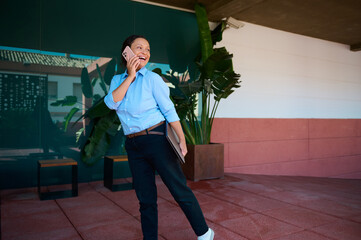 Smiling woman in casual attire talking on smartphone outdoors near office building with modern design and greenery