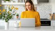 © Shisanupong - A woman in a yellow sweater working on her laptop in a bright kitchen with coffee and flowers, representing a home office setup