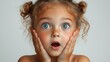 © KN Studio - Little Girl's Amazement: A close-up portrait of a young girl with bright blue eyes and a wide-open mouth, hands cupping her cheeks in astonishment. The image captures the innocence and wonder of child