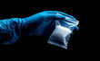 © Curioso.Photography - A gloved hand holds a small transparent bag containing white powder in a dark setting.