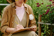 © AnnaStills - Person sitting in garden, writing notes on clipboard with yellow pencil Flowers blooming and greenery in background, creating serene outdoor atmosphere