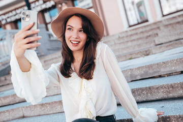 Wall Mural - Young beautiful smiling hipster woman in trendy summer white blouse and jeans. Carefree woman posing in the street in sunny day. Positive model outdoors. Cheerful and happy, takes selfie