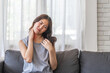 © Art_Photo - Asian woman sitting on sofa with a painful expression holding her neck, suffering from neck pain indoor home setting, casual clothing, woman health issue, discomfort, stress, fatigue at living room