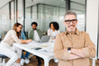 © Vadim Pastuh - Confident man with grey hair and glasses stands with his arms crossed, smiling at the camera, portraying leadership and experience, on background diverse group of professionals engaged in discussion