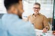 © Vadim Pastuh - A mature professional man in a tan shirt beams at a colleague out of focus in the foreground, exemplifying a supportive and collaborative office environment during a productive meeting.