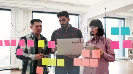 Wall Mural - Three colleagues collaborating on project using laptop and sticky notes. Team brainstorming ideas in modern office space. Capturing creativity, teamwork, and productivity in a professional setting.