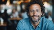 © Dulemegapixel - A cheerful middle-aged man with curly hair and glasses sits in a cozy cafe wearing a denim shirt, giving a warm and inviting smile amid a blurred warm background.
