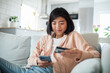 © Davor - Young Asian woman holding credit card and smartphone on home sofa
