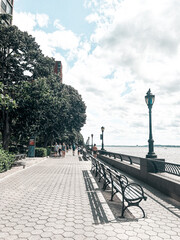 A scenic promenade with benches, street lamps, and trees along a waterfront. People stroll under partly cloudy skies, enjoying the peaceful setting