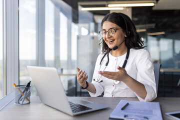 Wall Mural - A young female doctor with a headset talking to a patient, a clinic worker consults remotely, online support, an Indian woman uses a laptop, happily smiles and discusses the client's illness