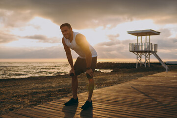 Wall Mural - Full body young strong sporty athletic toned fit sportsman man wearing sports clothes lean on knees warm up training at sunrise sun dawn over sea beach outdoor on pier seaside in summer day morning.