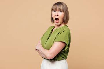 Side view elderly frowning sad blonde woman 50s years old wearing green shirt casual clothes hold hands crossed folded look camera isolated on plain light beige background studio. Lifestyle concept.