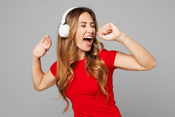 Young expressive singer happy woman wear red t-shirt casual clothes listen to music in headphones sing song in microphone at karaoke club isolated on plain grey background studio. Lifestyle concept.