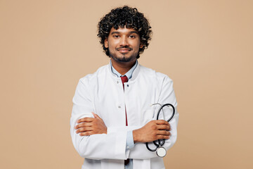 Wall Mural - Young confident Indian doctor man in white medical gown suit work in hospital clinic office hold in hand stethoscope look camera isolated on plain beige background studio Healthcare medicine concept