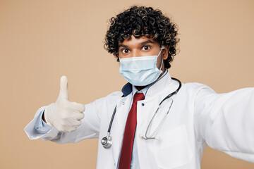 Wall Mural - Young Indian doctor man wear white medical gown suit mask work in hospital clinic office do mobile cell phone selfie shot show thumb up isolated on plain beige background. Healthcare medicine concept.