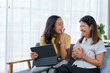© amnaj - Two women enjoying a light hearted moment on a tablet, giggling and chatting on a comfortable couch with tea, cultivating a warm and cheerful vibe in a cozy living space