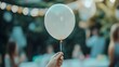 © sarana - Hand Holding a Balloon String: A hand holding a string attached to a floating balloon, with a soft-focus background of a birthday party.