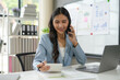© amnaj - Confident businesswoman multitasks in her office, calculating financial data and chatting on the phone, showcasing her skills in finance