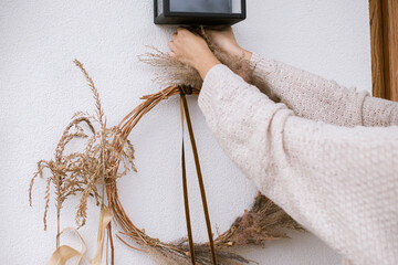 Wall Mural - Woman decorating house entrance with stylish autumn wreath. Rustic wreath with dried grass, herbs and ribbon in hands. Fall decor of front door and arrangement. Thanksgiving and Halloween