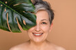 © InsideCreativeHouse - Closeup cropped portrait of a mature middle-aged woman with grey hair shirtless naked looking at camera covering her face with tropical leaf. Anti-age cosmetics for rejuvenation, anti-wrinkle effect