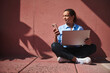 © Taras Grebinets - Confident woman sitting with laptop and smartphone against a colorful wall, working remotely and enjoying sunlight