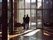 © Vitalii Shkurko - Two businessmen in suits having a conversation in a modern office lobby, with large windows letting in natural light and city buildings visible outside.