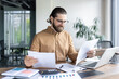 © Liubomir - Smiling businessman wearing eyeglasses analyzing reports at desk with laptop, papers, and calculator. Professional in modern office environment, focusing on data and strategy for business growth.