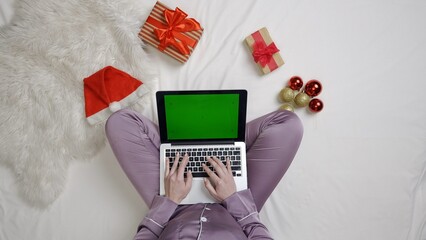 Wall Mural - Top view of female sits on the bed using gadget with chroma key green screen. Girl in pajama on the bed holding laptop typing text.