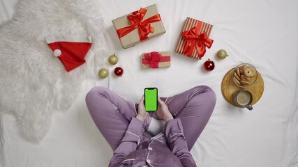 Wall Mural - Top view of girl on bed surrounded by gifts and snacks uses gadget chroma key green screen. Woman in pajama in bed holding smartphone.