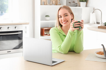 Wall Mural - Young woman with cup of hot tea and laptop sitting at table in kitchen