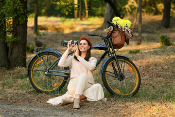 Wall Mural - Beautiful young happy woman with bicycle, camera, wicker basket of flowers and plaid sitting in park, outdoors