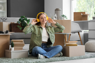 Wall Mural - Young woman in headphones with cardboard boxes sitting in room on moving day