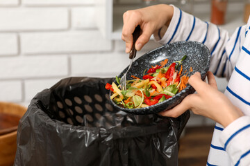 Young woman throwing organic food into trash bin in kitchen, closeup. Waste recycling concept