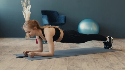 Sticker - A young woman holds a plank position on an exercise mat, focused on her phone. The room features fitness equipment, creating an active environment for her workout routine.