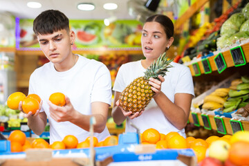 Male and female customers in casual clothes buying pineapples and oranges in grocery store