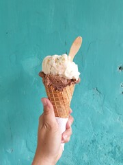 Hand holding corn ice cream with cheesecake and dark chocolate flavors on a turquoise background and a small spoon on top