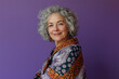 © Enrique - Portrait of a senior woman with curly hair wearing a patterned shawl, smiling at the camera, posing for a photo in the studio with lilac, purple background. international women's day